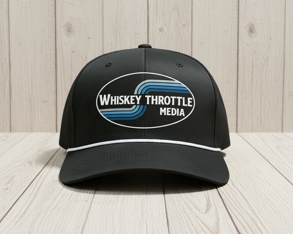 A black baseball cap with a curved brim, featuring a white oval patch on the front that reads 'Whiskey Throttle Media' with blue and black wavy stripes. A thin white piping detail runs along the edge of the brim.